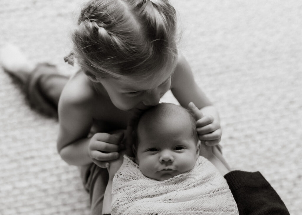 black and white portrait of newborn baby with older sister kissing his head